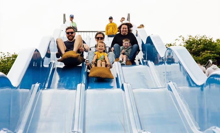 3 adults and 2 children sliding down a big blue slide in sacks.
